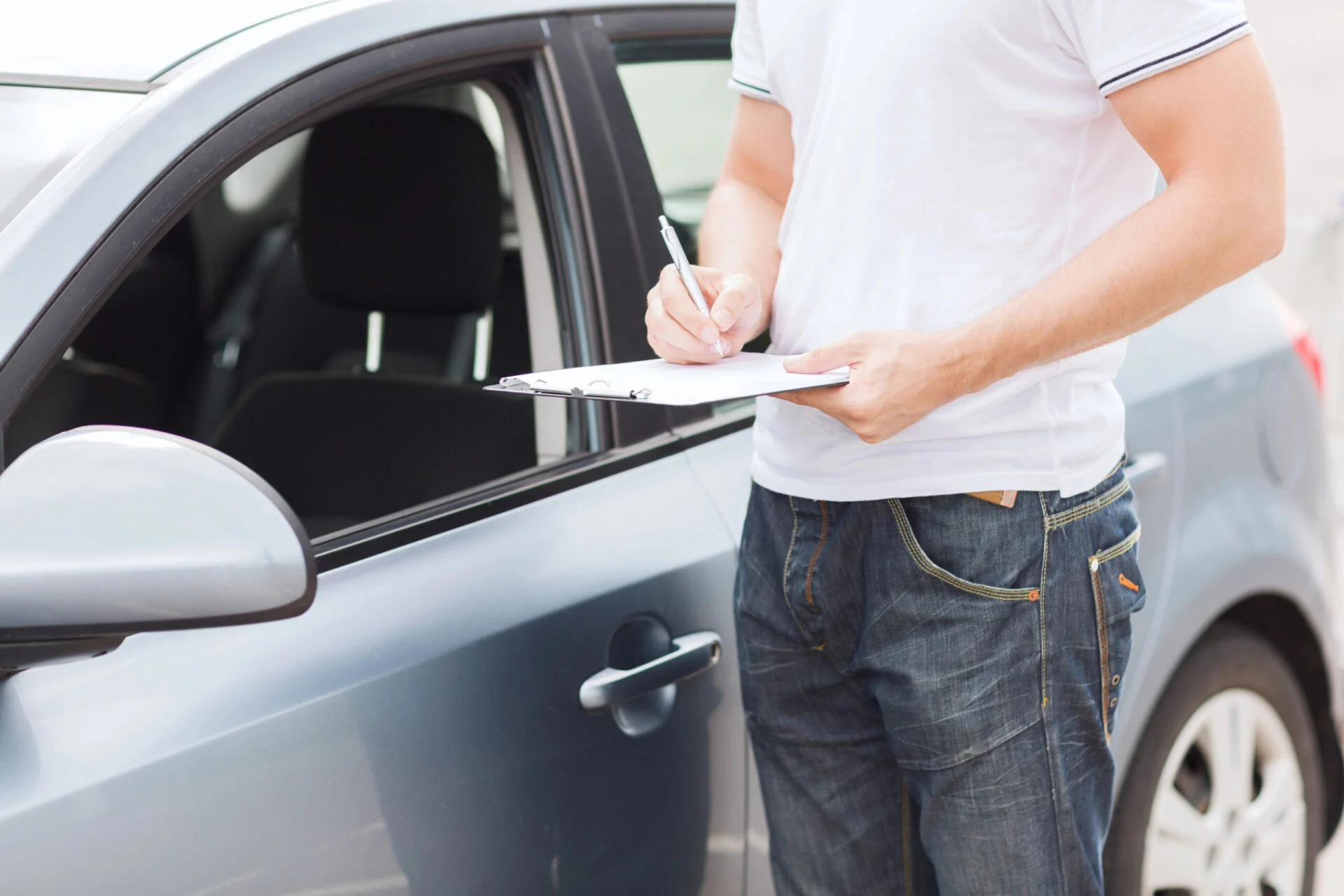 Man in white shirt holding a clipboard inspecting grey vehicle