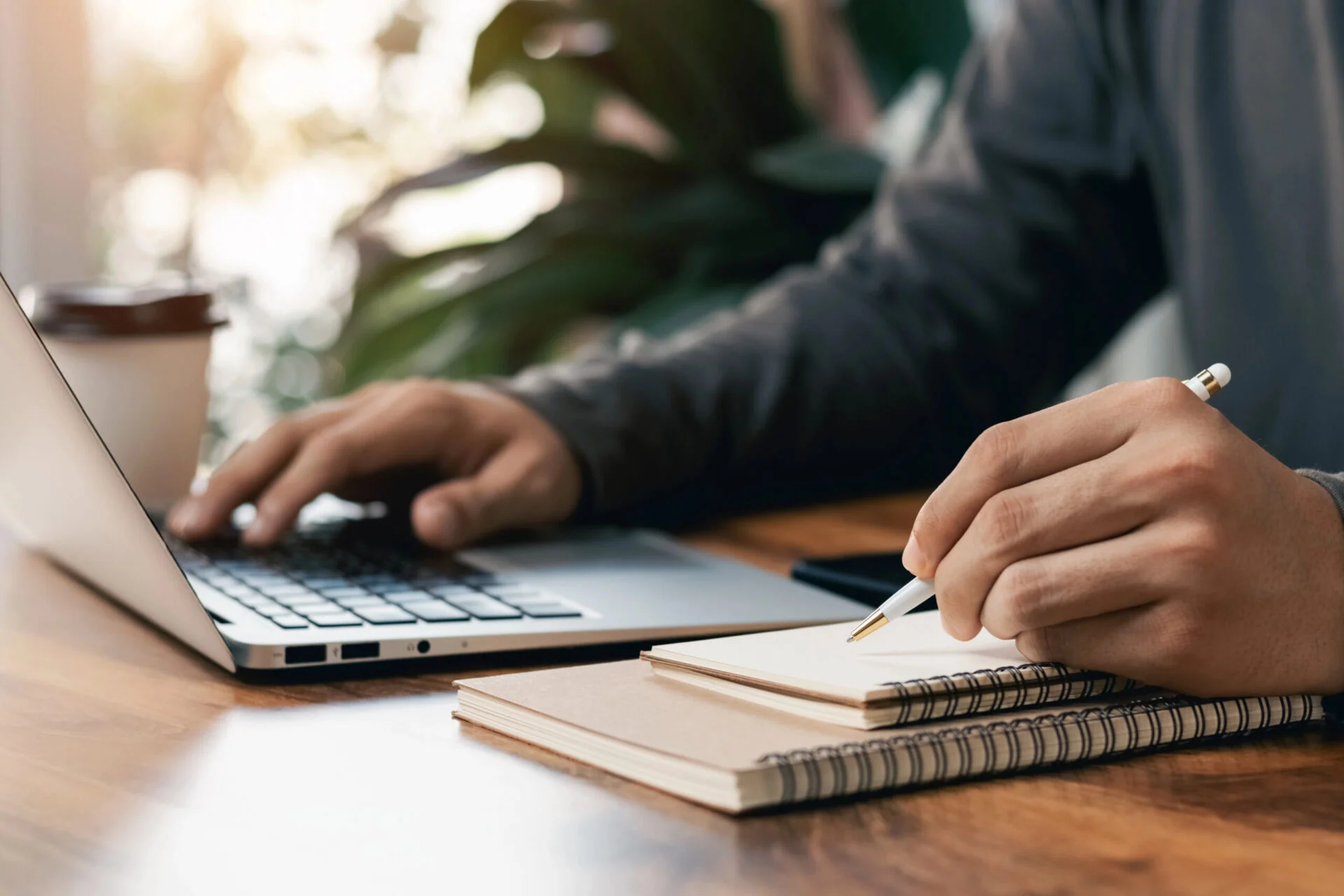 A mans hands typing on Macbook while writing on a notepad.