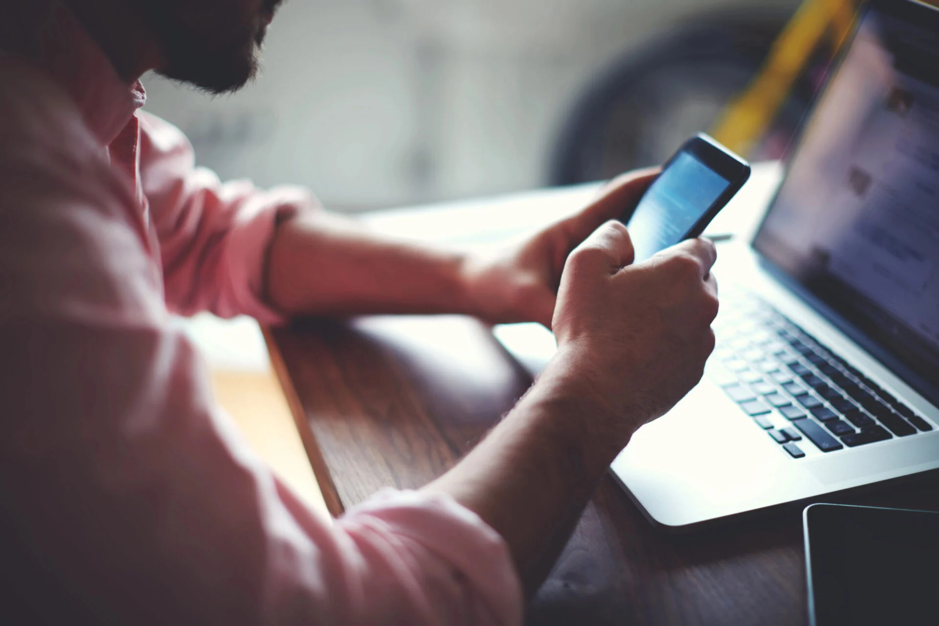 Person scrolling on a cell phone at a desk with a laptop in the background